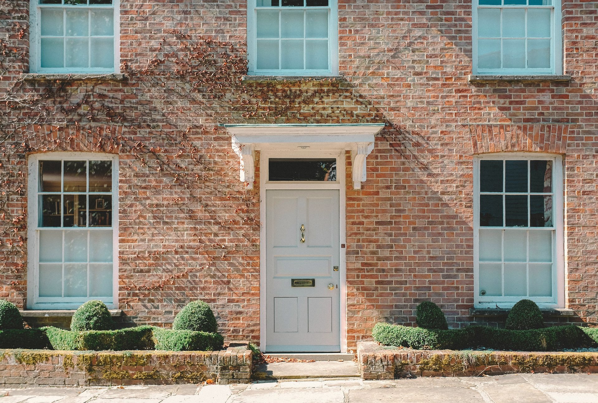 A white front door set in red brick, between tall sash windows and clipped boxwoods