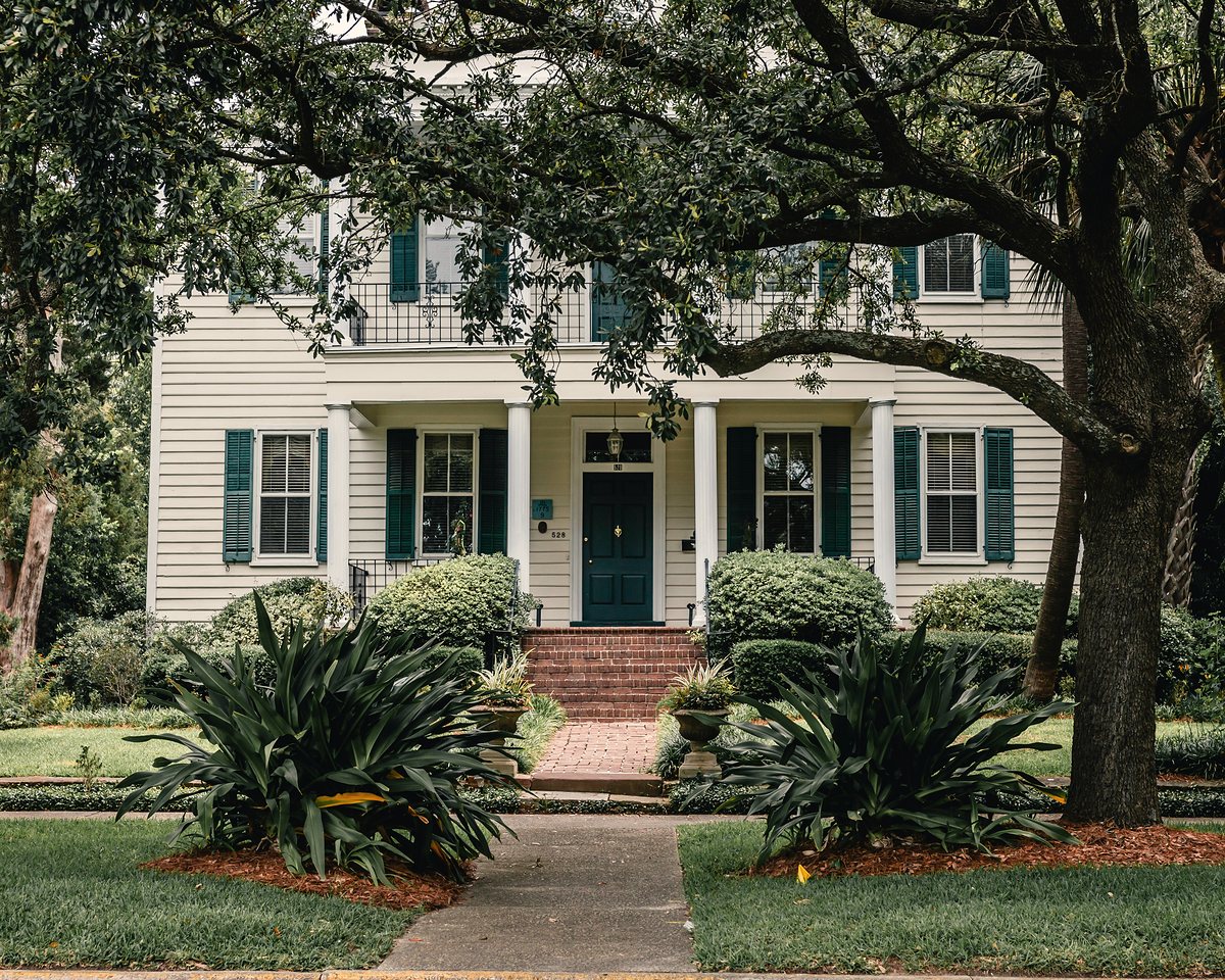 A white two story house with dark green shutters beneath a large live oak